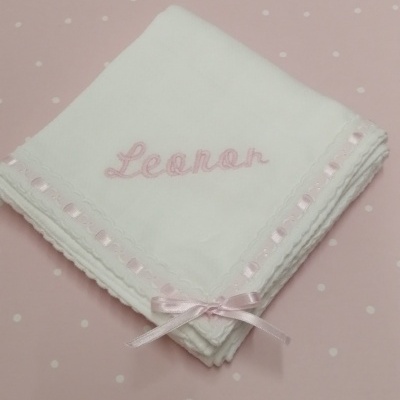 Fralda branca bordada com nome Leonor e fita rosa sobre fundo rosa com pontos brancos