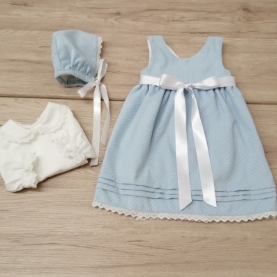 Conjunto de bebé com vestido azul claro, gorro e camisa branca sobre madeira clara