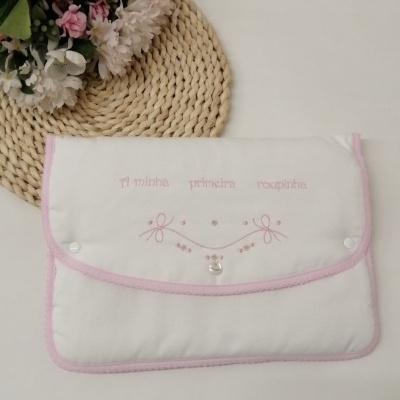 Envelope de tecido branco com bordas cor-de-rosa, bordado e texto rosa, sobre superfície branca ao lado de tapete de palha e flores.