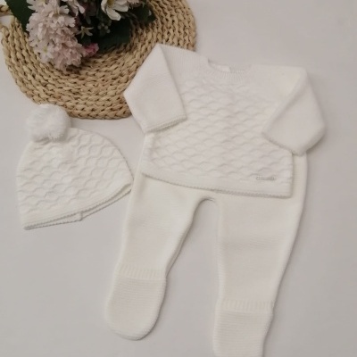 Conjunto branco de roupa de bebé com camisola, calças e gorro com pompom ao lado de flores sobre tapete de ráfia
