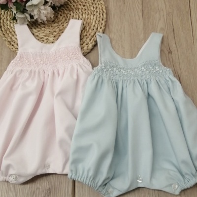 Dois macacões de bebé cor-de-rosa claro e azul claro com franzidos e botões, sobre chão de madeira e tapete de palha com flores.