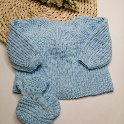 Conjunto de roupa de bebé azul clara em malha com camisola e meias, sobre fundo branco com base de palha e flores brancas.