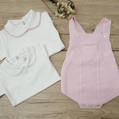 Conjunto de roupa de bebé com dois bodies brancos e uma jardineira rosa clara em malha sobre madeira.