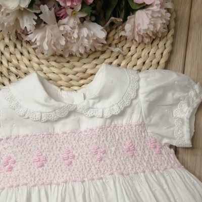 vestido branco de bebé com detalhes em renda e faixa rosa com padrão de ponto, colocado sobre tapete tecido e madeira com flores décor