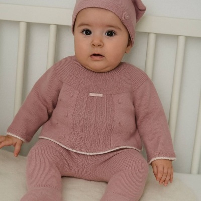 Bebé com roupa de malha rosa sentado numa cama branca.
