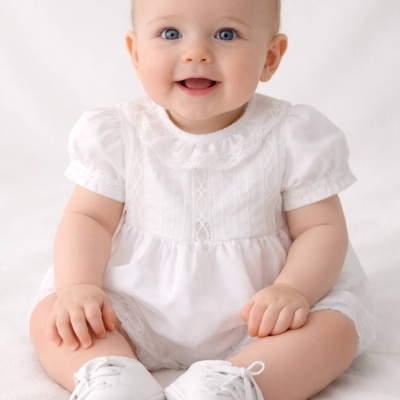 Bebé sentado com vestido branco com renda e sapatinhos brancos.