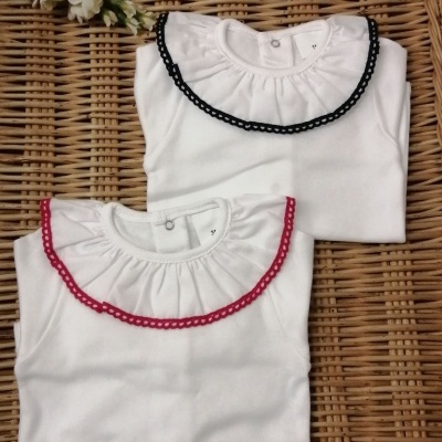 Dois bodies brancos de bebé com renda preta e vermelha no colarinho sobre superfície de vime com flores brancas