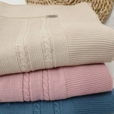 Três camisolas de malha dobradas em bege, rosa e azul com padrão trançado, sobre uma superfície branca com objeto de fibra natural ao fundo.