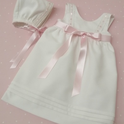 Vestido branco com fita rosa e boné branco com renda sobre fundo rosa com pontos brancos