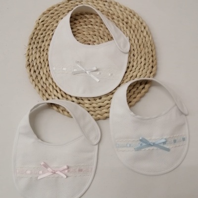 Três babetes brancos para bebé com laços de fita rosa, branco e azul e tapete redondo de fibras naturais