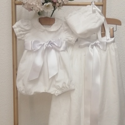 Dois conjuntos de roupa de bebé branca pendurados, um vestido curto com laço e um conjunto longo com fita e gorro, flores ao fundo.