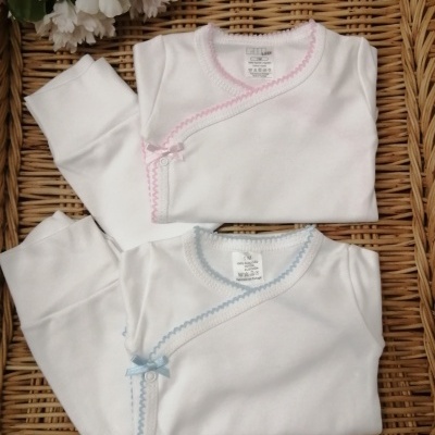 Dois conjuntos de roupa de bebé brancos com bordados rosa e azul sobre superfície de vime