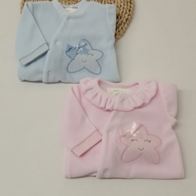 Dois macacões de bebé, azul e rosa, com estrelas e laços, sobre fundo branco com tapete redondo de palha.