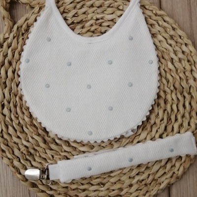 Babete e prendedor de chucha branco com pontos cinza sobre tapete redondo de fibras naturais.