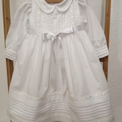 Vestido infantil branco com mangas compridas e laço frontal pendurado em cabide de madeira
