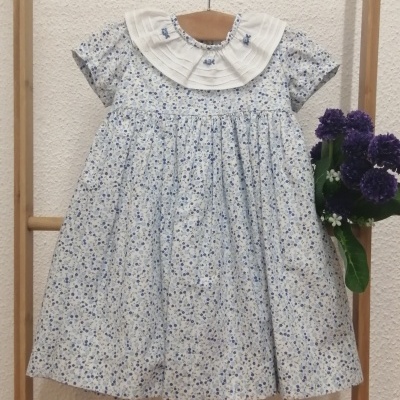 Vestido de bebé floral azul e branco com gola bordada, suspenso num cabide de madeira