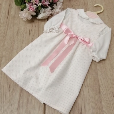 Vestido branco de bebé com laço rosa pendurado num cabide sobre madeira.