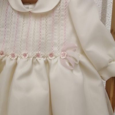 Vestido creme bebê com bordados rosas pendurado em cabide de madeira