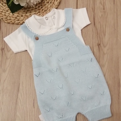 Macacão de malha azul clara com body branco sobre superfície de madeira clara e flores artificiais.