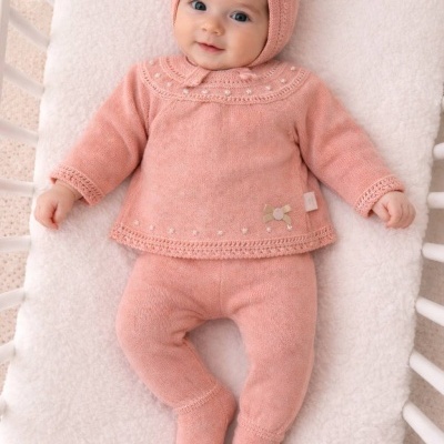 Bebé com conjunto de malha cor de rosa de manga comprida e gorro, deitada num berço branco com almofada felpuda branca.