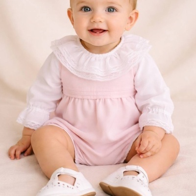 Bebé vestido com body rosa e branco com gola de renda e sapatinhos brancos sentado em fundo bege.