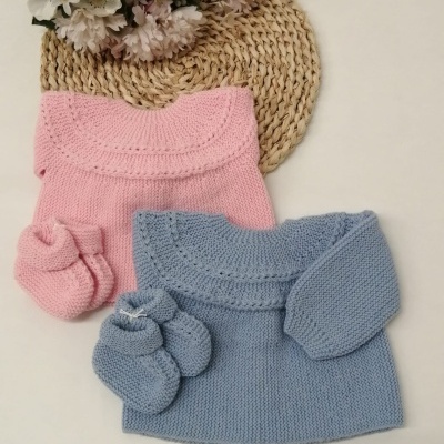 Dois conjuntos de roupa de bebé em malha rosa e azul com botinhas colocados sobre base redonda de fibra natural e flores artificiais