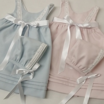 Dois conjuntos de roupa de bebé azul e rosa com vestidos e gorros, decorados com renda e laços brancos