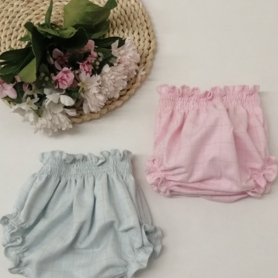 Dois calções curtos para bebé em azul claro e rosa com padrão em quadrados, elástico franzido, junto a suporte redondo de palha com flores artificiais