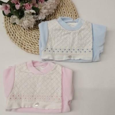 Conjuntos de roupa para bebé com camisola branca bordada e partes em tecido azul e rosa sobre mesa branca com tapete de palha e flores.