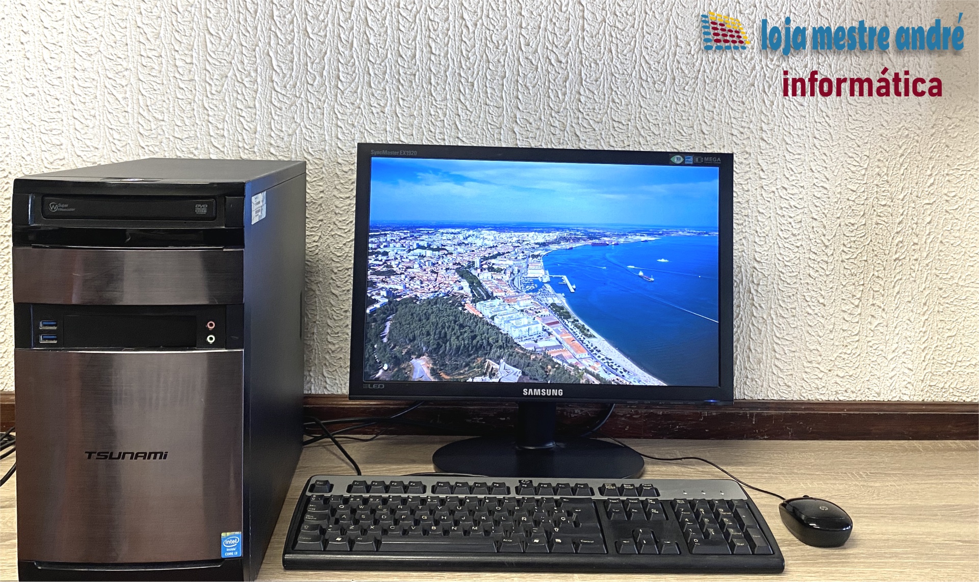TSUNAMI Computador de secretária com torre Tsunami e monitor Samsung, teclado e rato em mesa de madeira frente a parede texturada.