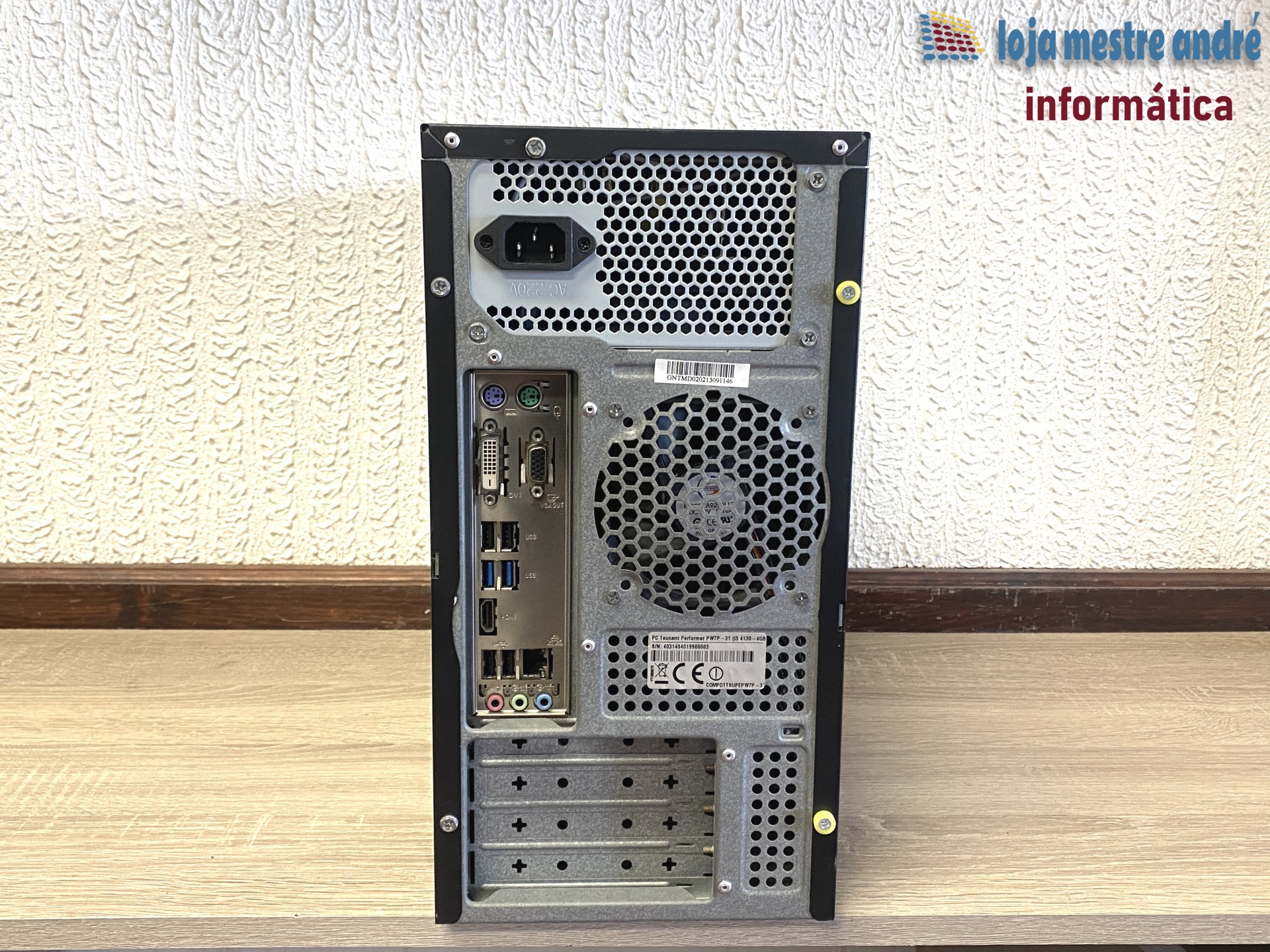 TSUNAMI Computador de secretária preto e cinza visto de trás com portas e ventoinha, sobre mesa de madeira clara e parede texturizada de fundo.