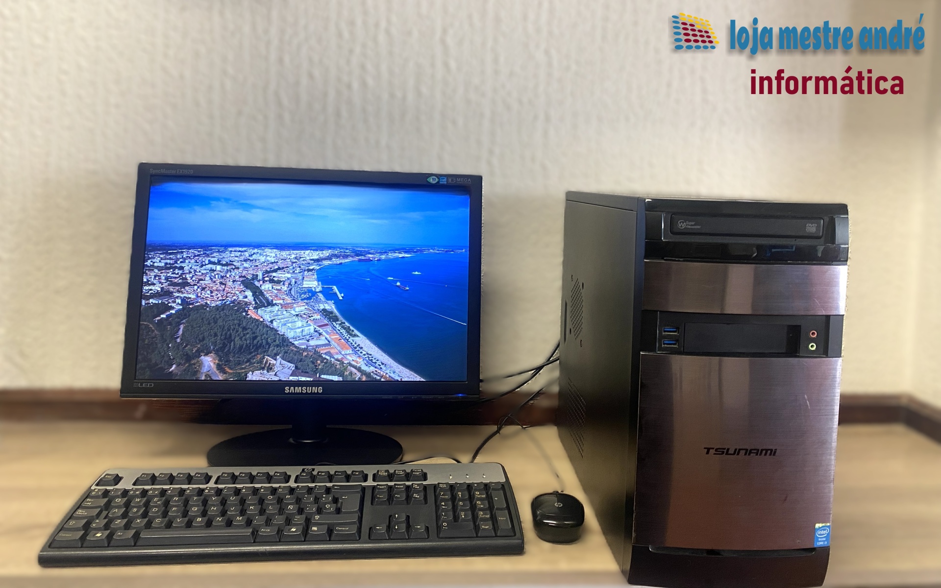TSUNAMI Computador desktop com monitor Samsung, teclado, rato e torre T-WORKS numa mesa com parede branca e texto da loja informática
