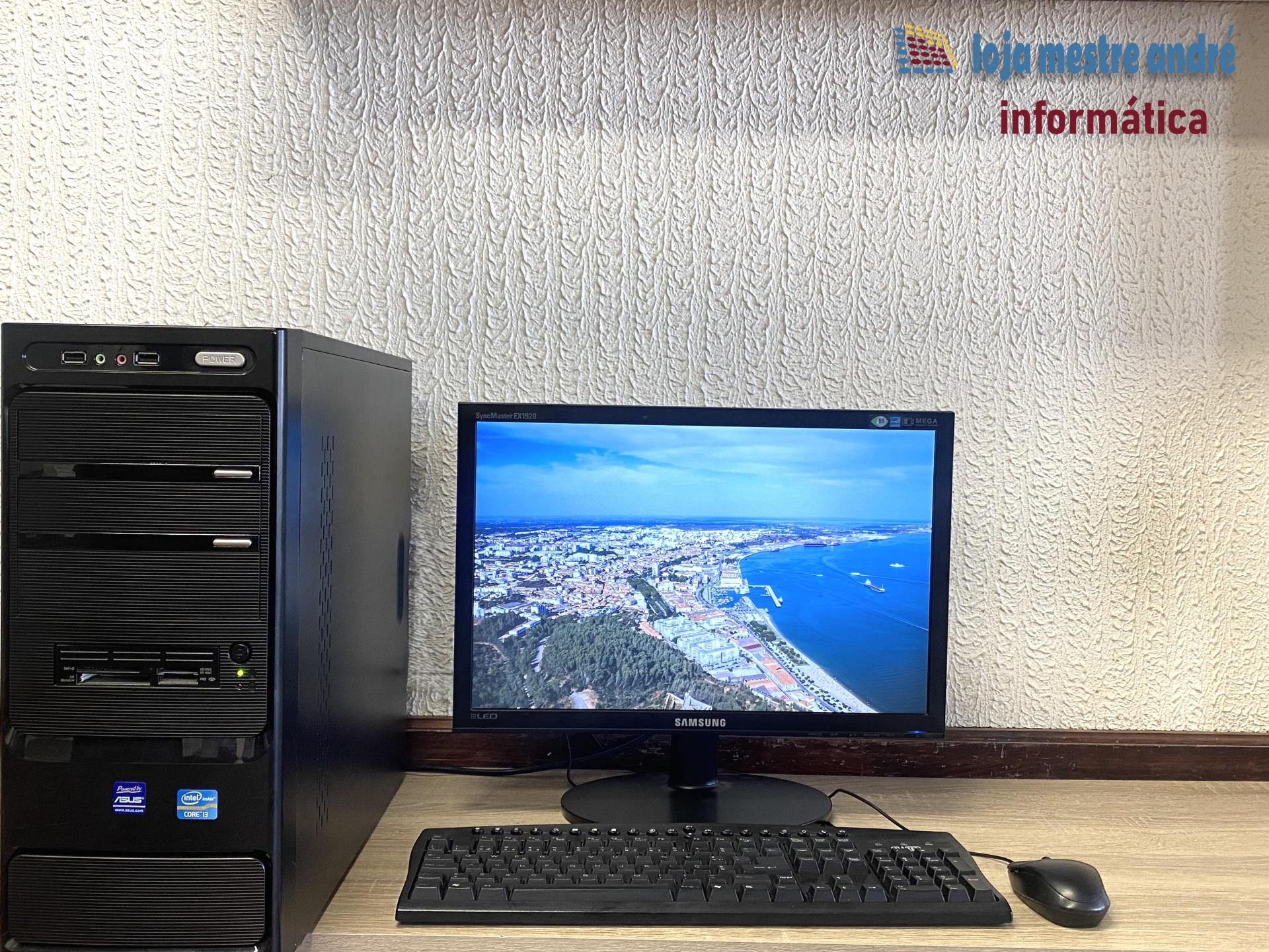 LMA B2 Computador desktop preto com monitor Samsung, teclado e rato na mesa, parede texturizada ao fundo com logotipo e texto