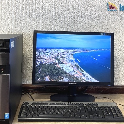 Computador de secretária com torre Tsunami e monitor Samsung, teclado e rato em mesa de madeira frente a parede texturada.