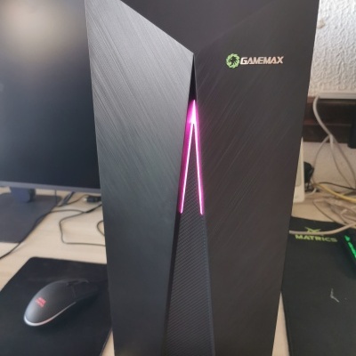 Caixa de PC gaming preta com luz LED roxa e logo GameMax, numa secretária com teclado, rato e monitor.