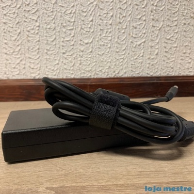 Carregador preto com cabo enrolado sobre chão de madeira clara junto a parede branca