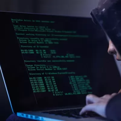 Portátil com ecrã de texto verde sobre fundo preto com comandos de sistema, usado por pessoa com capuz