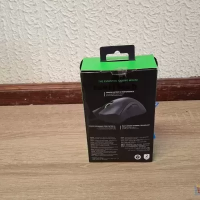 Caixa de rato gaming preta e verde em chão de madeira com parede branca texturada.