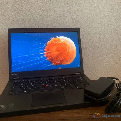 Portátil Lenovo preto com imagem de água-viva laranja no ecrã e carregador ao lado sobre madeira.