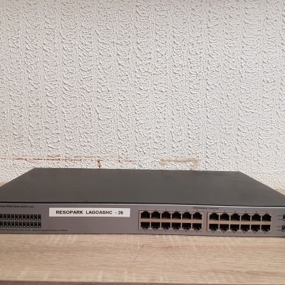 Comutador de rede Cisco de 24 portas preto e cinza numa superfície de madeira com parede branca texturizada ao fundo