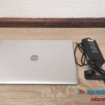 Computador portátil cinza prateado Dell com carregador preto sobre mesa de madeira sobre fundo branco texturado