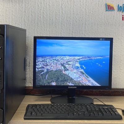 Computador de secretária preto com teclado, rato e monitor Samsung LED a mostrar imagem aérea do litoral.