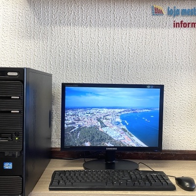 Computador desktop preto com monitor Samsung, teclado e rato na mesa, parede texturizada ao fundo com logotipo e texto