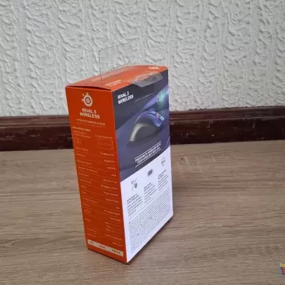 Caixa laranja de produto eletrônico SteelSeries Rival 110 sobre chão de madeira próximo a parede branca texturizada