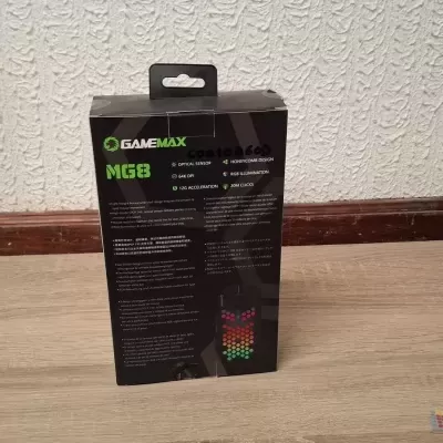 Caixa preta do GAMEMAX MG8 com texto em inglês e imagem colorida no chão de madeira clara contra parede branca