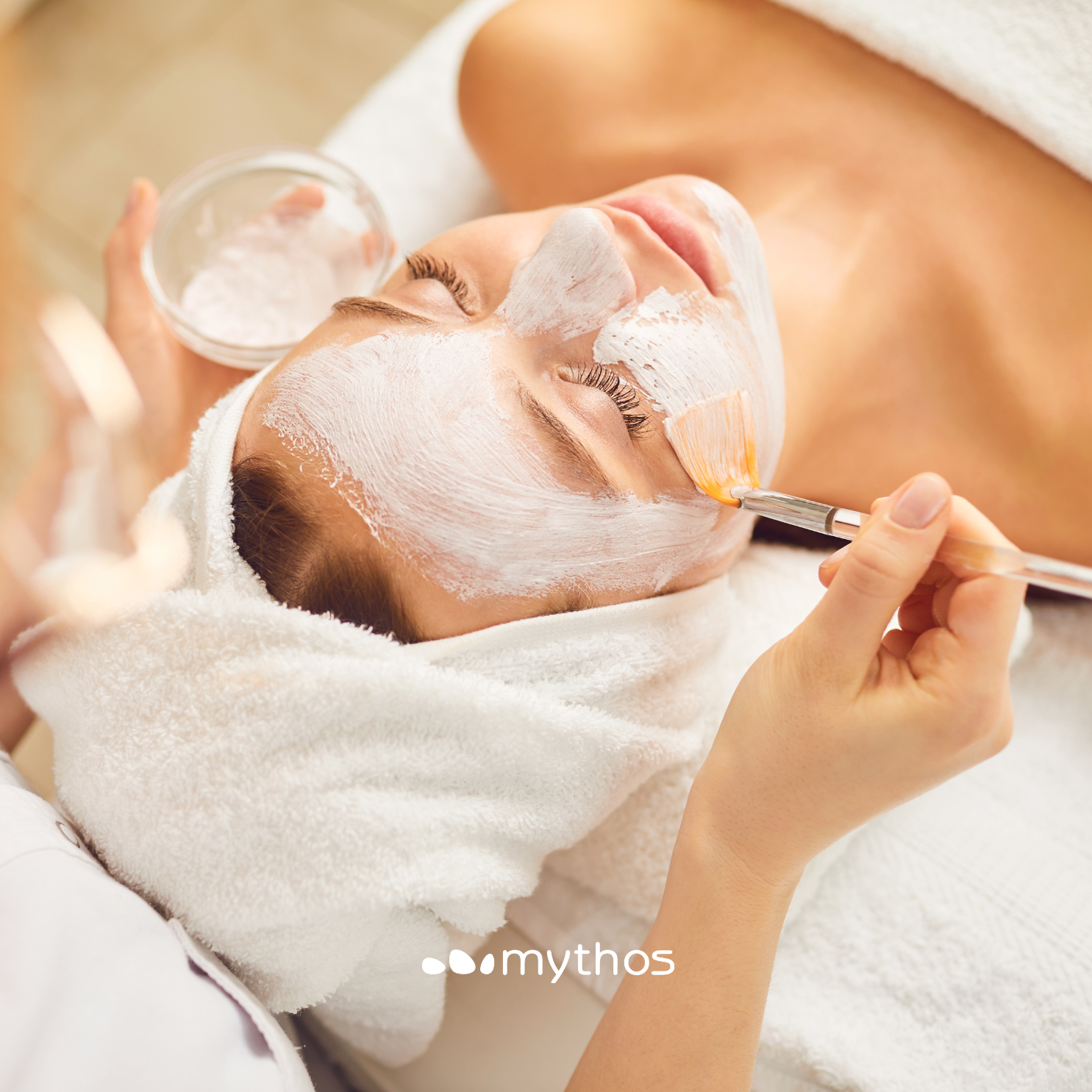 Limpeza de Pele Pessoa a receber aplicação de máscara facial branca com pincel, cabeça com toalha branca em ambiente de spa