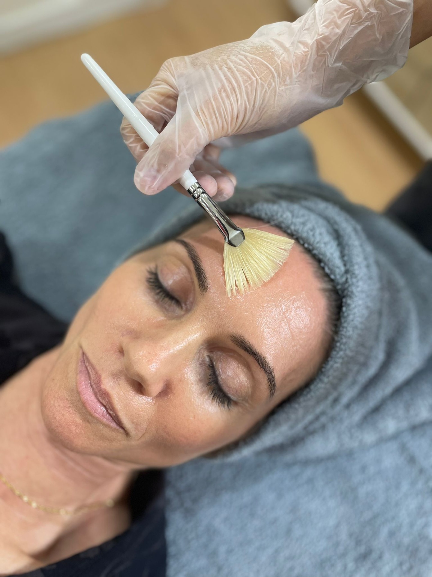 Peeling Quimico Mulher com toalha cinzenta na cabeça recebe aplicação facial com pincel branco