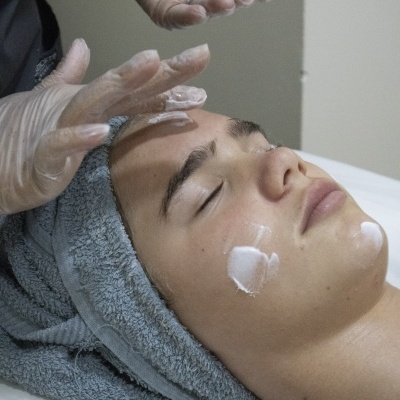 Pessoa a receber aplicação de creme no rosto com toalha na cabeça e luvas transparentes.