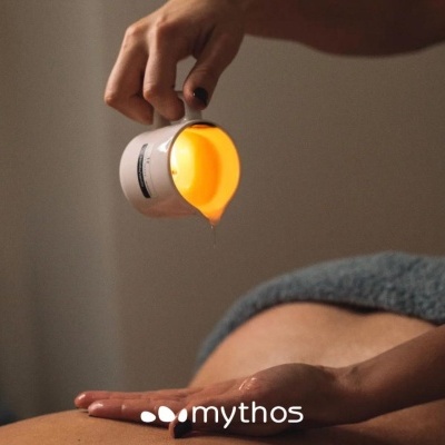 Mão a verter óleo dourado de recipiente branco sobre pele de pessoa deitada com toalha cinzenta