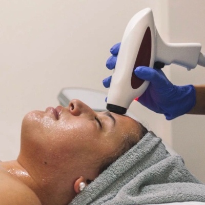 Pessoa a receber tratamento facial com aparelho branco e vermelho e toalha cinza na cabeça